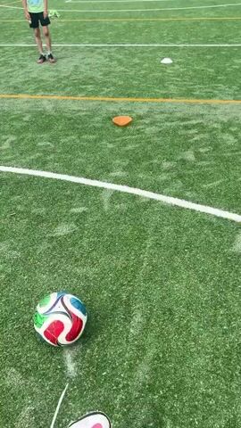 A boy wearing a lime green t-shirt and black shorts kicks a soccer ball on an artificial turf field. There are several small cones positioned on the field. The boy kicks the ball towards a soccer goal. The soccer field is enclosed by a green fence. Behind the fence is a building with a red roof. A basketball hoop is attached to a pole next to the soccer goal. In the background, other buildings and trees can be seen. The sky is overcast.