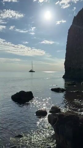 A serene coastal landscape is captured, featuring a large catamaran sailing or anchored on calm waters under a bright, clear sky. The sun shines intensely, creating sparkling reflections on the water in the foreground, where several dark rocks are partially submerged. A towering, textured cliff or rock formation occupies the right side of the frame, providing a dramatic contrast to the open sea. The sky is blue with scattered white clouds, suggesting pleasant weather. A voice speaking Russian is heard in the background. The scene appears peaceful and is located in Divnomorskoe, Russia.