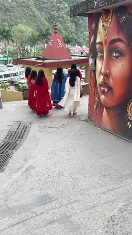 The video shows a group of six women walking on a paved road towards a red temple. The women are wearing traditional Indian clothing in various colors: orange, red, blue, and white. Next to the road is a building with a large mural of a woman's face, adorned with traditional jewelry.  In the background, there are mountains, trees, and a parking lot with several vehicles. A motorcycle passes by between the group of women and the mural. The camera remains static throughout the video.