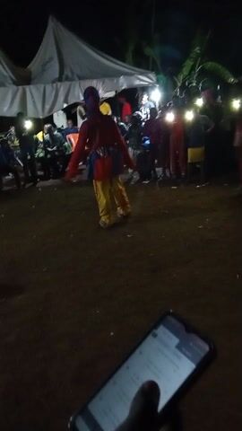 The video captures a nighttime scene of what appears to be a cultural performance or celebration. Several individuals in colorful costumes are dancing in a circle on a grassy area. They are surrounded by a crowd of spectators, some holding up lights, possibly phones, to illuminate the dancers. In the background, there is a large white tent and some banana trees. In the foreground, a smartphone is visible, suggesting that someone is recording the event.