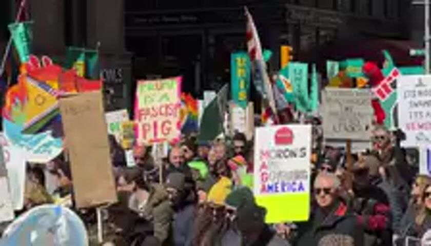 The video captures a large, dense crowd of people participating in an outdoor gathering, likely a protest or rally. Individuals are packed closely together, many holding various signs and flags. A prominent American flag, featuring the red and white stripes and blue field, is waving in the center of the frame throughout the video.

Many attendees are dressed in winter clothing, including coats, jackets, and some wearing hats and sunglasses. The atmosphere suggests an active demonstration, with numerous signs raised high.

Several signs have partially readable text:
- On the left side of the frame, a sign is partially visible with the text "...PIG".
- In the center-right, a bright yellow sign prominently displays the word "AMERICA" with other text above it that is difficult to discern.
- Another notable sign, seen clearly towards the right, carries the message "HATE WON'T MAKE AMERICA GREAT."
- Other signs are present, some with green or colorful backgrounds, but their text is mostly illegible due to blurriness or distance. One sign on the far left appears to have rainbow colors, and another near the "HATE" sign seems to show the number "7."

The background consists of a dark building, suggesting an urban environment, possibly a city street or plaza. The lighting indicates it is daytime. The crowd appears to be stationary or moving slowly, focused on displaying their messages through signs and flags.