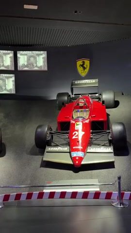The video shows a slow pan of several red Formula 1 cars displayed in a museum. The cars are in pristine condition and appear to be vintage models. The walls behind the cars are dark and feature framed black and white photographs of racing events and drivers. The Ferrari logo, a prancing horse, is visible on the wall. The cars are roped off with red and white striped barriers. The lighting is dim, with spotlights illuminating the vehicles. The overall atmosphere is one of reverence and historical appreciation for the sport.