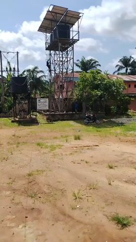 The video shows a tall, metal water tower with a dark container at the top, located on a grassy, sandy patch of land. There is a person climbing the tower. There is another smaller water tank on a shorter metal stand to the left of the taller tower. Nearby, there is a building partly obscured by trees. A parked motorcycle stands to the right of the tower. Power lines and poles are visible in the periphery. The sky is blue with scattered clouds.