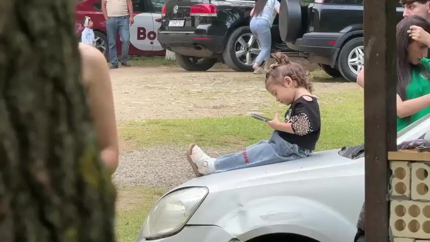 A group of people are gathered in an outdoor setting, likely a picnic or a social gathering. In the foreground, a young girl with dark hair tied in a ponytail is sitting on the hood of a white car, engrossed in a smartphone. She is wearing a black top with embellishments and blue jeans. To her right, a young woman in a green shirt is partially visible, interacting with something just outside the frame. To the left and slightly behind the girl, a woman with dark hair is also visible. In the background, several vehicles are parked, including a black SUV and a red car. A small child in a light-colored outfit stands near the red car. The ground is covered with gravel and grass. The overall atmosphere suggests a casual, informal gathering.