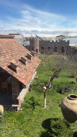 The video shows an outdoor courtyard or garden area on a sunny day. There's a pavilion-like structure with a red-tiled roof and brick pillars, containing wooden tables and chairs. The ground is covered in grass with yellow dandelions. Several large clay pots are visible, some near a small stone patio area. In the background, there's a brick wall and a building resembling a small castle tower. Beyond that, greenhouses are visible. The camera pans across the scene, providing a wide view of the courtyard.