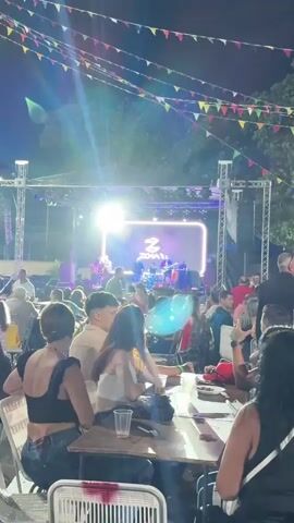 The video shows an outdoor event taking place at night. A band called Zona 2 is playing on a stage, illuminated by stage lights and backed by a large screen displaying the band’s logo. A crowd of people is seated at tables, enjoying the music while drinking from plastic cups and eating. Colorful triangular flags are strung above the tables, adding to the festive atmosphere. The setting appears to be a patio or open space, with trees visible in the background. People are dressed casually, and some are looking at their phones while others watch the band. One person wearing a red dress dances in the foreground.