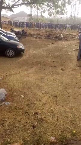 The video shows a large group of people gathered in an open, dirt-covered area. Several cars and motorcycles are parked to the left. A man in a grey outfit is standing to the right, holding what appears to be a camera or recording device. The ground is uneven, with small rocks and sparse vegetation. There is a tree in the background, and the scene is filmed on a sunny day.