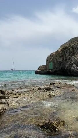 This is a vertical, eye-level shot of a rocky coastline on a bright, partly cloudy day. The foreground features wet, textured rocks with clear, shallow water lapping over them. In the midground, the turquoise sea stretches to the horizon, with a white sailboat visible in the distance on the left. To the right, a rugged, rocky cliff rises, with a small, green-painted doorway set into its base. A stone staircase leads up to the doorway from the water's edge. Small waves are breaking against the rocks in the midground. The overall impression is of a tranquil, secluded coastal scene.
