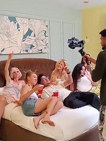 A man films a group of six people on a bed. Five of the people on the bed are women, and one is a man lying on his back. The women are sitting or lying around him, some holding red plastic cups. The cameraman holds a camera mounted on a stabilizing rig. The room is decorated in light colors with a large painting above the bed. The people on the bed are looking at the camera and smiling.