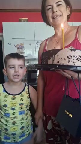 A woman and a young boy are standing in what appears to be a kitchen. The woman is holding a chocolate cake with a lit candle on it. She is smiling at the camera. The boy is standing next to her, looking somewhat pensive. The woman is wearing a pink tank top and patterned pajama bottoms. The boy is wearing a yellow tank top with cartoon characters on it. There is a blue gift bag on the right side of the frame. The background shows kitchen cabinets and a counter. The overall impression is a birthday celebration.