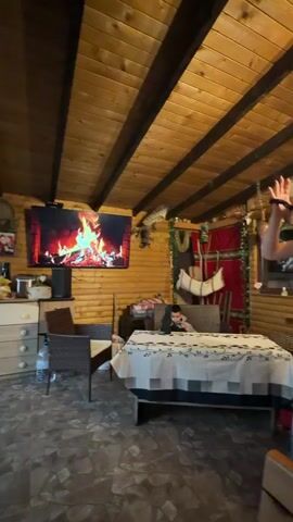 A woman and a man are dancing in a cabin, holding hands. The woman is wearing a light green shirt and light purple pants, while the man is wearing a blue shirt. In the background, a young boy is sitting at a table, looking at a tablet. The cabin has wooden walls and a wooden ceiling, with a TV showing a fireplace scene on the wall. A bottle of water sits on the floor near a chair. The cabin is decorated with rugs, pillows, and other decor. The couple appears happy and carefree as they dance.