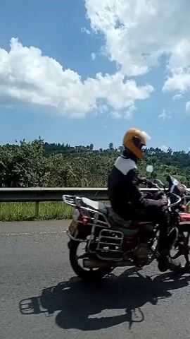 The video shows a view from a moving vehicle, driving along a road with a metal guardrail. The landscape beyond the guardrail features lush, green vegetation covering rolling hills. The sky above is bright blue, dotted with fluffy white clouds.