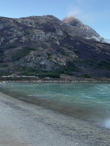 The video shows a scenic landscape featuring a partially frozen lake against a backdrop of mountains. The water is a light turquoise color with thin ice sheets on the surface. The shoreline is a gravelly beach. Mountains rise sharply behind the lake, exhibiting rocky terrain and sparse vegetation. Some snow is visible on the mountain peaks. The sky is a pale blue, suggesting either early morning or late afternoon light. No people or animals are visible in the scene.