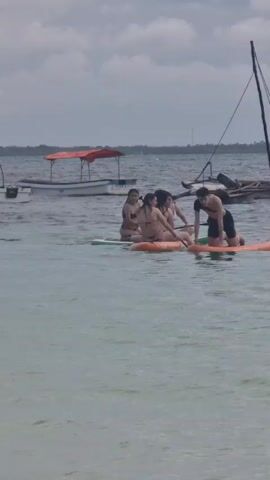 Four women wearing bikinis and one man wearing shorts are on two connected paddleboards in the ocean. The paddleboards are orange and green. The man is kneeling on the orange paddleboard. One of the women is sitting on the orange paddleboard. Two women are sitting on the green paddleboard. One woman on the green paddleboard is using a paddle. A child is swimming in the water near the paddleboards. In the background are several boats, a cloudy sky, and a distant shoreline.
