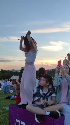 A young woman with long, curly red hair is dancing on a purple platform outdoors. She is wearing a white crop top and loose pink pants. In the foreground, a young boy wearing a black baseball cap and a plaid shirt draped over his shoulders sits on the edge of the platform. To the right of the boy, a young woman is holding up a pink smartphone, seemingly recording the dancer. Several other people are visible in the background, some sitting on the grass, and others standing. The sky is a pale blue with wispy clouds, suggesting it is either early morning or late afternoon. The overall setting appears to be a park or open field where an event is taking place.