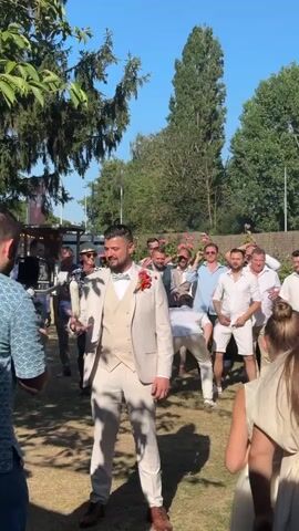 A man in a light-colored suit with a bowtie and a boutonniere is holding a white scroll in his right hand and is mid-motion, appearing to be either throwing or unfurling it. He is surrounded by a group of men, most of whom are wearing white shirts and shorts, and a few women. The setting appears to be an outdoor gathering, possibly a wedding reception, with greenery, trees, and a clear blue sky visible. The men are engaged in activity, with one man in the background bending over. The overall atmosphere seems festive.