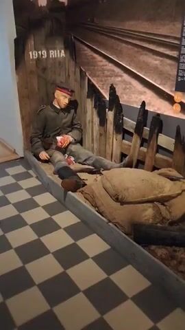 The video shows a diorama of a wounded soldier sitting in a trench. The soldier is wearing a military uniform and has injuries to his hand and leg. A rifle is lying next to him. The trench is made of wood and sandbags, and there is a checkered floor in front of it. A sign above the soldier reads "1919 RIIA".