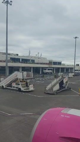 The video shows an airport tarmac on an overcast day. Two sets of mobile stairs are positioned on the tarmac, with a white and grey airport building in the background. A white bus is parked near the building. An airport worker in a yellow vest walks across the tarmac, pulling a piece of equipment.  In the lower right corner, part of the pink engine of an airplane with the words "NO STEP" is visible.