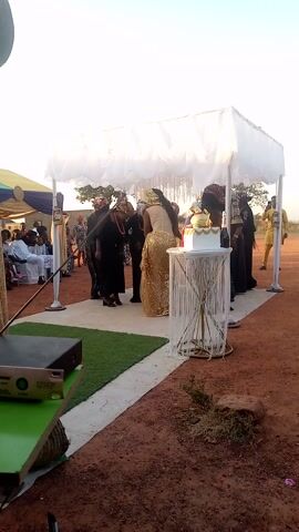 This video captures an outdoor celebratory event in Unguwan Doma, Nigeria, under a bright, clear sky. The scene is set on reddish-brown dirt ground, typical of the region.

A prominent white, rectangular canopy structure serves as the focal point, adorned with white sheer fabric. Underneath this canopy, a group of people, primarily women, are gathered. A woman wearing a shimmering golden, long-sleeved dress is seen embracing another woman dressed in black. Other women in dark attire are also present, appearing to dance or sway to music, creating a festive atmosphere. A decorated white table with intricate white and gold accents stands under the canopy.

Leading away from the canopy is a white path or carpet, bordered by a strip of green artificial grass. Further in the background, additional guests are seated on white chairs under another canopy, indicating a larger gathering. A person in a yellow garment is visible standing further back in the distance.

In the immediate foreground, to the bottom right, audio equipment, possibly a speaker or mixer, is visible on a green surface, with partial text "WH..." discernable on its front panel. The bright daylight suggests the event is taking place during the day. The overall mood is joyful and celebratory, with people engaged in dancing and social interaction, characteristic of a wedding or similar community event.