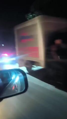 The video shows a nighttime scene from inside a vehicle. The camera is looking out the side window and reflects the interior of the car in the side mirror. In the distance, cars and trucks are visible on a road, their headlights and taillights creating streaks of light due to motion blur. The environment appears to be urban or suburban, with buildings and trees visible along the roadside. The road is paved and appears to be in use by multiple vehicles. The overall impression is a sense of movement through a nighttime cityscape.
