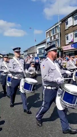 A marching band parades down a street on a sunny day. The band members are dressed in matching light gray shirts, dark blue pants, and dark caps. Many are playing instruments, including drums and what appear to be brass instruments like trumpets or cornets. The drummers are carrying snare drums. The band is moving from left to right in the frame. In the background, there are buildings lining the street, and some spectators can be seen on the sidewalk. The sky is blue with scattered white clouds.