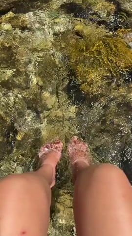 This is a first-person perspective video of someone's legs and feet wading through shallow, clear water with a rocky bottom. The water is moving, creating ripples and distortions on the surface. Sunlight illuminates the water and rocks, showing green and brown hues. The person's bare feet are visible, with their toes occasionally disappearing beneath the water's surface. One leg has a small red mark or abrasion on the knee. The focus is on the interaction between the person's legs and feet and the natural environment of the water and rocks.