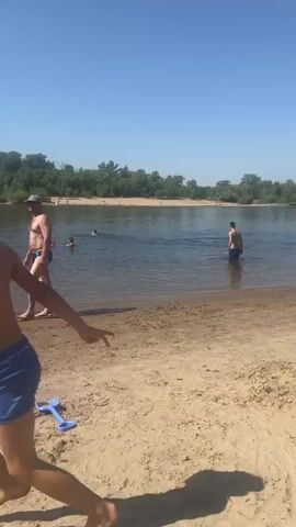 A man and a young boy are on a sandy beach next to a body of water. The man is wearing swim trunks and a hat, walking along the shoreline. The boy is also in swim trunks, and he is running towards the water. There are two toy trucks and a blue shovel on the sand. In the water, several people are swimming. The sky is clear and blue. The background is filled with trees.