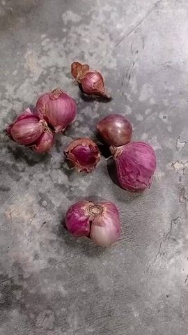A top-down shot shows a cluster of red onions on a textured grey surface. The onions are of various sizes, with some appearing as single bulbs and others clustered together. Their papery outer skins are a deep reddish-purple, with lighter pinkish-white layers visible where they are slightly peeled or damaged. One onion in the foreground has a distinctive, almost heart-like shape. The background surface appears to be concrete or a similar rough material, with slight variations in color and texture. There are no people or actions depicted in the video, it is a static image with subtle lighting shifts.