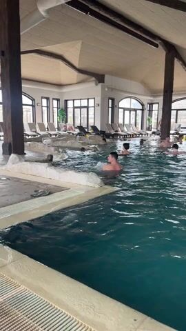 The video shows an indoor swimming pool with several people in it. The pool is a dark teal color and has several sections with different depths. There is a large seating area along the side of the pool with lounge chairs. The building has high ceilings with exposed wooden beams. Large windows let in natural light. The camera moves around the pool, showing different angles and people enjoying the water.