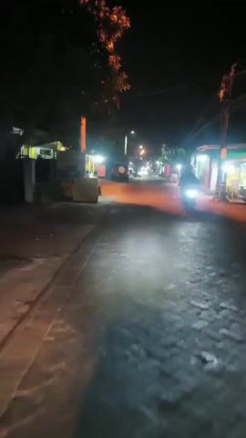 A nighttime street scene with several buildings and vehicles visible. The road is lit by streetlights and the headlights of approaching vehicles. There are several food stalls or small shops lining the street, with some displaying illuminated signs. A dark-colored SUV with its trunk open is parked on the left side of the road, and a person is standing near it. Further down the street, other vehicles are visible, and some people appear to be walking or standing outside the establishments. The overall impression is of a lively street, possibly a market area, operating after dark.