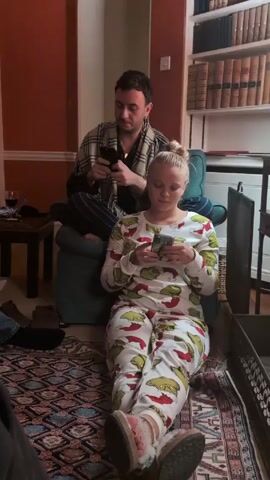 In a residential living room, a young woman sits cross-legged on a patterned rug wearing Grinch-themed pajamas and furry slippers, looking at her phone. Behind her, a man in a plaid robe and striped pants sits in an armchair, also looking at his phone. A small wooden table with a wine glass is to the left, and a bookcase filled with books is on the right.