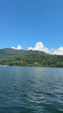 The video shows a body of water with a wooded hill in the background. The sky is blue with scattered white clouds. On the water, a yellow boat is moving from left to right. There are a few scattered buildings visible on the hillside. The water is choppy, suggesting movement or wind. The overall scene depicts a natural landscape with some human presence.