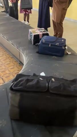 The vertical video shows a baggage carousel with a few pieces of luggage moving around the bend. Three adults stand near the carousel, their bodies partially visible from the knees down. One person on the left wears a white and red patterned dress and white sneakers. The person in the middle wears a dark blue dress and dark shoes. The person on the right wears brown pants and a patterned shirt, holding a dark bag. The carousel is dark grey with some lighter markings. The floor is light brown with a patterned design. The wall in the background is light yellow.