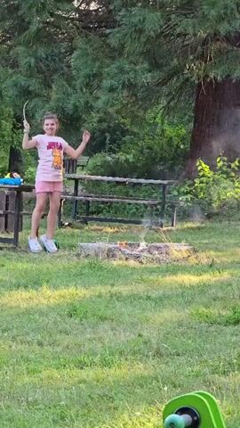 A young girl in a pink t-shirt and shorts is standing in a grassy area next to a campfire. She is holding a stick and appears to be poking the fire. There are trees and picnic tables in the background.