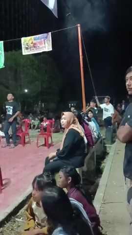 The video captures a nighttime outdoor scene, possibly a sporting event or gathering, with a volleyball court visible in the background. Several people are present, some seated on red chairs, others standing or sitting on the ground. A woman wearing a hijab is prominently seated in the foreground, facing left. In the foreground, a group of young people are also seated, with one pointing towards the left side of the frame. The court is illuminated by artificial lights, creating shadows. Banners are hanging from poles above the court, suggesting an organized event. The overall atmosphere appears casual and communal.