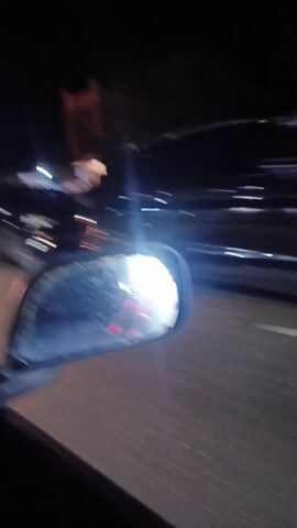 The video shows the view from the side mirror of a vehicle driving at night. A variety of vehicles pass by on the right side of the road, including a black truck, a white van, and a pick-up truck with people in the back under a blue canopy. Streetlights and illuminated businesses are visible along the side of the road. The side mirror is dirty.