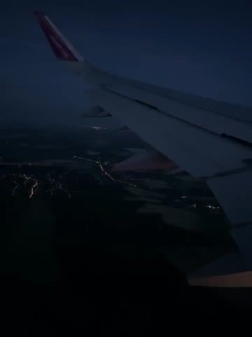 The video captures a view from an airplane window at night. The prominent subject is the airplane's wing, occupying a significant portion of the right side of the frame. The wing is white, with a distinctive pink/purple wingtip visible towards the left, where the Wizz Air logo is faintly discernible. Below the wing, a dark landscape stretches out, illuminated by numerous clusters of artificial lights from towns, roads, and possibly larger settlements. These lights appear as bright, scattered points and lines against the dark ground, indicating populated areas. The sky above is a dark blue, suggesting twilight or night. The airplane appears to be in flight, likely during ascent or descent, providing an aerial perspective of the illuminated terrain below. No people are visible in the video. The scene depicts a calm night flight over what appears to be a rural to semi-urban landscape. The location context provided is Gomba, Hungary, which aligns with the view of dispersed lights over a varied terrain at night.
