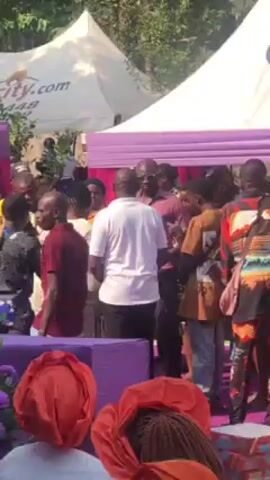 The video shows a group of people gathered near a tent. The tent has a white top and purple trim. A woman in a cream-colored lace dress is visible in the foreground. A man in a white shirt stands next to her. Several other people stand around them, including a person wearing a Michael Jordan jersey. The people appear to be interacting with each other, possibly celebrating something. The location seems to be outdoors, with trees and other vegetation in the background. A sign with "City.com" is visible on one of the tents.