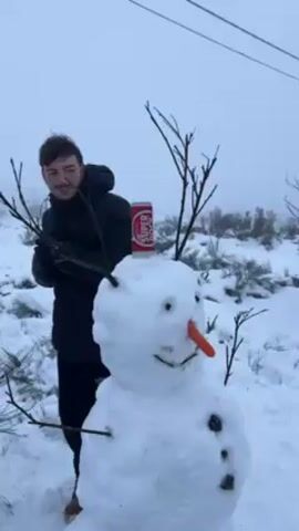 The video shows a man standing in front of a snowman in a snowy environment. The snowman has a carrot nose, twig arms, and a can of Super Bock beer on its head. The man smiles, adjusts the can, and then walks away from the snowman. The background includes snowy terrain and scattered bushes. Above, power lines are visible. The weather is overcast.