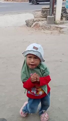 A young child wearing a white hat and a green headscarf is standing in a dirt area. The child has on a red long-sleeved shirt with a cartoon image on it and blue pants. The child's hands are clasped together and then move outwards as if clapping. The child then looks directly at the camera and smiles. In the background, a road with a vehicle on it is visible.