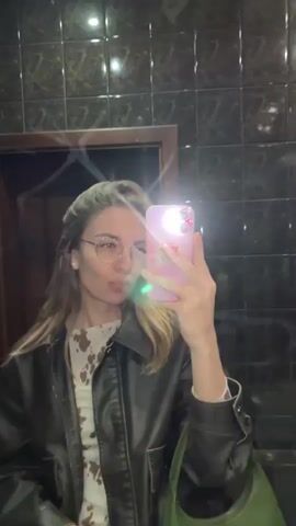 A woman is taking a selfie in a tiled bathroom with her pink phone. She is wearing glasses, a brown leather jacket over a patterned shirt, and has a green purse over her shoulder. She begins by blowing a kiss at the camera, then stops. The bathroom has dark tiles and a wooden door.