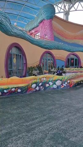 The video shows an outdoor scene under a large, open-air canopy structure. The architecture is colorful and whimsical, with undulating, wave-like designs and circular windows. The foreground is a paved area with a textured, mottled gray surface. To the left, there's a decorative low wall with sculpted shells, coral, and fish. Behind this, the building has a curved facade adorned with a vibrant, multi-colored mural that resembles ocean waves and sand. There are several entrances with blue doors and purple circular frames.

In the mid-ground, three people are walking away from the camera, towards the building. Two are in the foreground, closer to the camera, and the third is slightly behind them. To the left, a person is sitting on a bench next to a lamppost. Further into the scene, more people are visible walking and congregating near the building entrances. The background shows more of the colorful, themed architecture, including structures that resemble large, colorful fish or sea creatures, and a central dome-like structure. The overall impression is of an amusement park or entertainment venue with a strong aquatic theme. The lighting suggests it might be late afternoon or early evening.