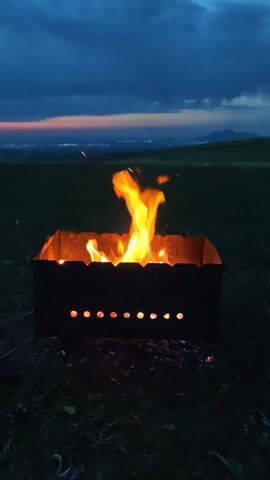 A metal barbecue grill stands on a grassy field, with flames burning inside. The grill is black with several circular holes along the bottom. The flames are yellow and orange, reaching high into the air. The background is a twilight sky with a reddish-orange horizon and dark blue clouds. In the distance, a city skyline can be seen. The ground around the grill is covered in green grass.