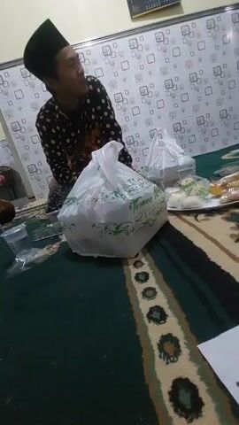 A person wearing a black peci and a black and white patterned shirt is sitting on a green carpet. They are looking to their right. In front of them are two white plastic bags with green designs. To the right of the bags is a plate with some food on it, including what appears to be a yellow item and some white items. There is also a clear plastic cup with liquid in it to the left of the bags. The background shows a wall with a checkered pattern. Another person is partially visible on the far left of the image. The lighting appears to be artificial.