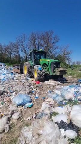 The video shows a green John Deere tractor parked amidst a large pile of discarded plastic bags, bottles, and other trash in a sunny outdoor area. The camera pans slowly to the left, revealing the extent of the garbage pile against a clear blue sky and trees with bare branches in the background. The ground is dry and cracked, with litter scattered throughout. The tractor's tires are muddy. There are no people visible in the shot.