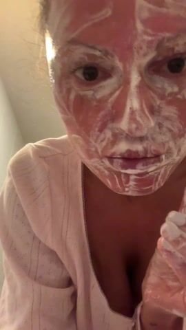 A close-up shot captures a person applying a white, foamy substance to their face with both hands. The person's face is covered in the substance, with their eyes visible. Their lips are slightly puckered, and they appear to be concentrating on the task. The individual's clothing suggests they are indoors, possibly in a bathroom. Water droplets are visible on their hands.