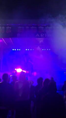 A male rapper is on stage in front of a large crowd, performing into a microphone. He is wearing a black cap, t-shirt, vest, and shorts. The stage is illuminated with blue and white spotlights, and a backdrop with text in a foreign language is visible behind him. A haze hangs in the air. The crowd is mostly obscured, with several people holding up their phones to record the performance. The rapper gestures with his hands as he performs.