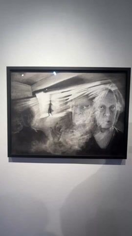 The video shows a framed black and white artwork hanging on a white wall. The artwork depicts what appears to be a hallway or room with light coming from a doorway. A shadowy figure is walking towards the light. Overlaid on the hallway scene are two faces. The faces are slightly blurred, giving a ghostly or dreamlike effect. The artwork is in a black frame. The camera zooms in on the artwork. Background sounds suggest there are people talking nearby.