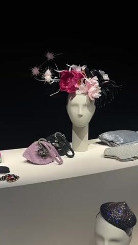 The video displays a fashion exhibition. The first shot shows a mannequin head adorned with a flamboyant hat featuring pink and white flowers and black feathers. Several small handbags are arranged on a white shelf in front of it. To the right, there are two sequined clutches. The camera then pans down to reveal another mannequin head wearing a black hat covered in sparkling rhinestones. More items, including shoes and clutches, are displayed on a lower shelf. The final shots show a wider view of the exhibition space with various accessories, including more shoes, handbags, and jewelry, presented on white shelves against a black background.