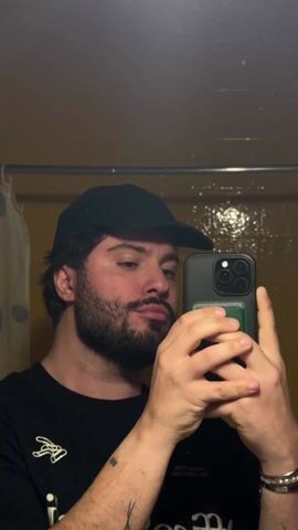 A person wearing a black cap and black t-shirt stands in front of a mirror and holds a phone to take a selfie. The person has a beard and dark hair, and they stick their tongue out. The background includes a yellow tiled wall and a shower curtain.